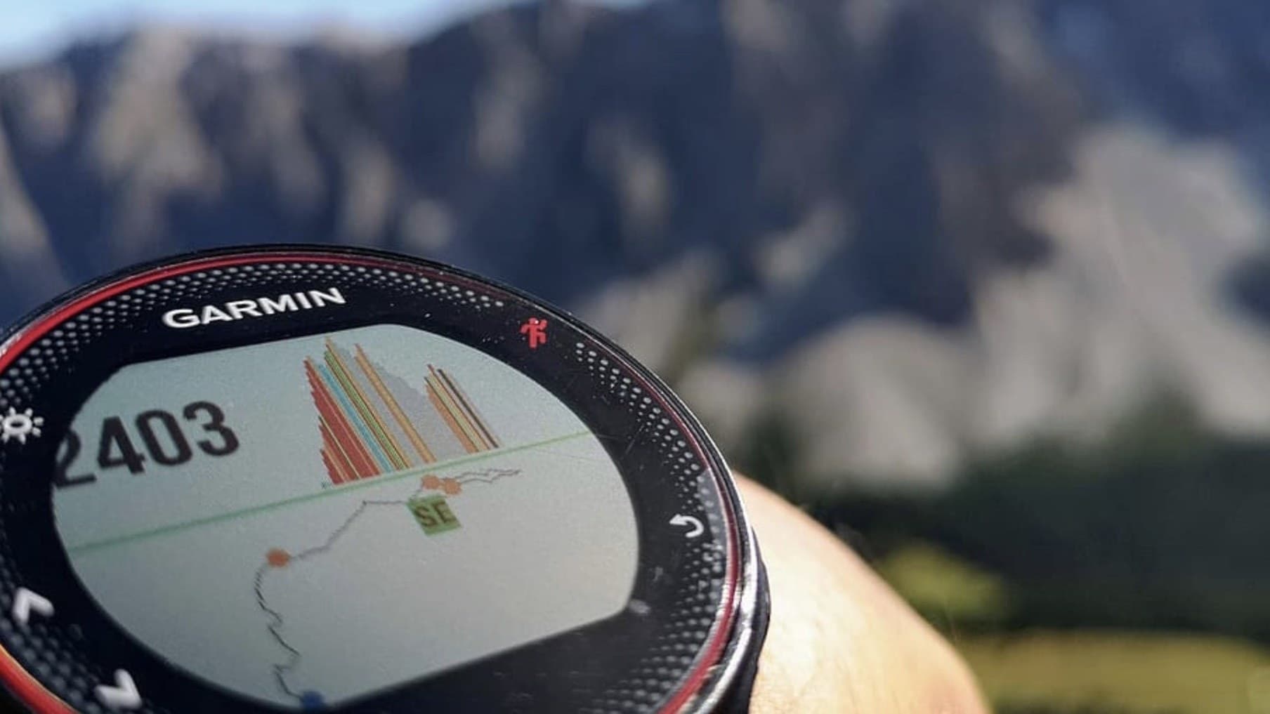 Garmin watch on a wrist showing a loaded course with waypoints and an elevation profile, mountains behind