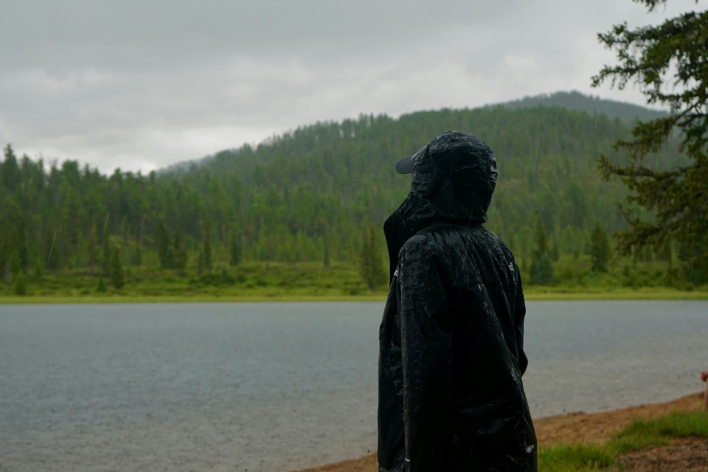 Hiker in a wet hooded rain jacket by a lake in steady rain, illustrating waterproof ratings explained
