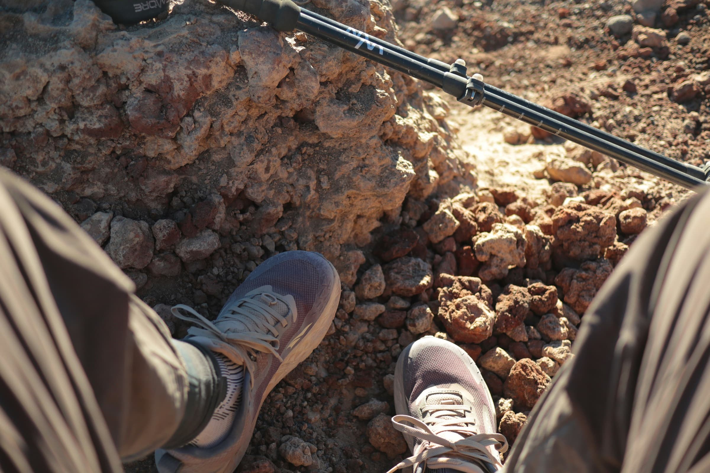 Hiker's view down at hiking shoes on rocky red ground with a trekking pole, comparing Salomon vs Merrell